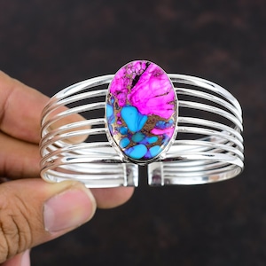 May include: A silver bangle bracelet featuring a multi-coloured oval gemstone. The gemstone is a mix of pink, blue, and gold colours.
