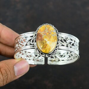 May include: A silver cuff bracelet with an oval-shaped yellow, brown, and white gemstone set in the center. The bracelet has a delicate, floral design.