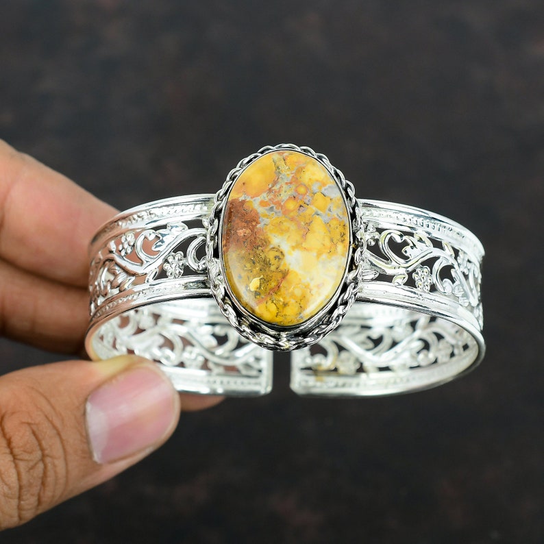 May include: A silver cuff bracelet with an oval-shaped yellow, brown, and white gemstone set in the center. The bracelet has a delicate, floral design.