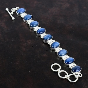 Könnte beinhalten: Ein silbernes Armband mit einem Verschluss verfügt über eine Reihe von ovalen, blauen Edelsteinen. Das Armband ist aus Sterlingsilber gefertigt und hat eine polierte Oberfläche.