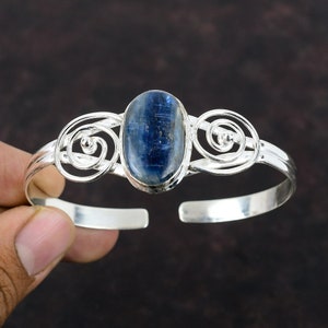 Könnte beinhalten: Ein silbernes Armreif-Armband mit einem blauen Edelstein, der in einem Wirbelmuster eingefasst ist.
