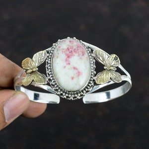 May include: A silver cuff bracelet with a large oval, cream-colored stone speckled with red. The stone is set in a silver frame with a beaded border. Gold-colored butterfly accents are on each side of the stone.