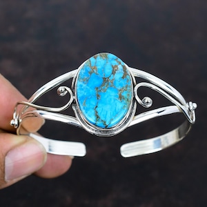 May include: A silver cuff bracelet featuring an oval turquoise stone with brown matrix patterns. The bracelet has an open design with decorative swirls and is held by a hand against a dark background.