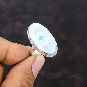 Könnte beinhalten: Ein Silberring mit einem großen, ovalen Larimarstein. Der Stein hat eine hellblaue Farbe mit weißen und dunkelblauen Mustern. Der Ring wird von einer Hand vor einem dunkelbraunen Hintergrund gehalten.