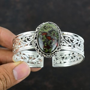 May include: Silver cuff bracelet featuring an oval-shaped stone with green and red hues. The stone is set in a decorative silver frame with a chain-like border. The bracelet has an open design with intricate floral cutouts.