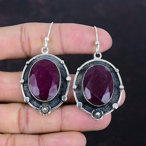 May include: A pair of silver drop earrings. Each earring showcases a large, oval, deep red gemstone set in a decorative frame. The frame has a dark, textured border with small, round accents and a floral detail at the base.