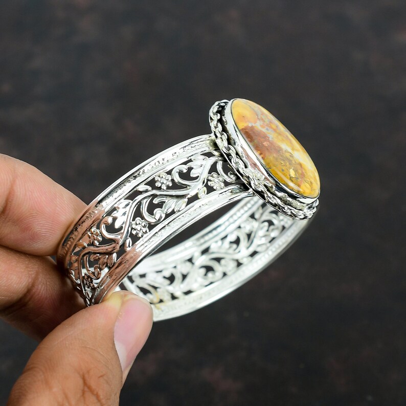 May include: Silver bangle bracelet with an oval-shaped, brown and yellow agate gemstone set in a silver bezel. The bracelet has a delicate, floral design.