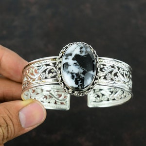May include: A silver cuff bracelet with an oval-shaped black and white gemstone set in a silver bezel. The bracelet has a delicate floral design.