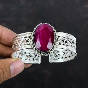 Könnte beinhalten: Silbernes Armreif-Armband mit einem ovalen, tiefroten Edelstein. Das Armband hat ein offenes Design mit aufwendigen floralen Filigranmustern. Der Edelstein ist in eine silberne Lünette gefasst.