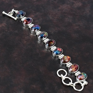 May include: A silver bracelet featuring oval-shaped gemstones with a mix of colors, including blue, red, and purple. The bracelet has a toggle clasp and is displayed on a dark brown surface. The gemstones are set in silver bezels.