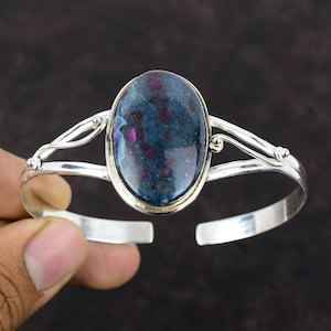 May include: A silver bangle bracelet with a large oval-shaped blue and pink gemstone set in the center. The gemstone has a speckled pattern.