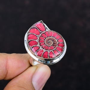 Op de afbeelding: Een zilveren ring met een gepolijst ammonietfossiel met een levendig rood en bruin spiraalpatroon. De zetting van de ring is helder zilver, wat de ingewikkelde details van het fossiel benadrukt. De ring wordt tegen een donkere achtergrond gehouden.