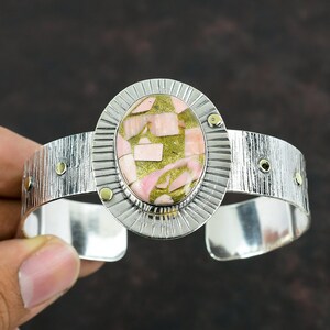 May include: A silver cuff bracelet with a pink and green gemstone set in a silver and gold bezel. The bracelet has a textured surface and is adjustable.