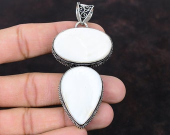 May include: A handcrafted pendant featuring two white, oval-shaped stones. The stones are set in a silver-colored metal with a decorative border. The pendant has a decorative bail at the top for a chain. The pendant is held in a hand.
