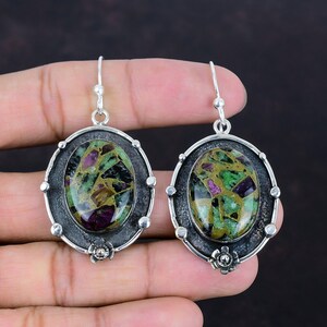 May include: A pair of silver earrings with oval-shaped green, black, and pink gemstone settings. The earrings have a floral design at the bottom.