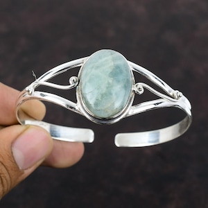 Puede incluir: Un brazalete de plata con una piedra ovalada de color azul claro engastada en el centro. El brazalete tiene un diseño delicado y ornamentado con dos bandas curvas.
