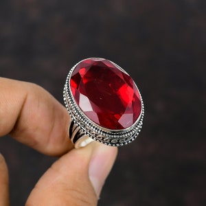Op de afbeelding: Een ring met een grote, ovaal geslepen, rode edelsteen en een sierlijke zilveren band. De edelsteen is gefacetteerd en vangt het licht. De ring wordt tegen een donkere achtergrond gehouden.