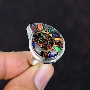 Può includere: Un anello in argento con un fossile di ammonite lucido e iridescente come elemento centrale. Il fossile presenta un motivo a spirale con vivaci riflessi multicolori di rosso, verde, blu e arancione su uno sfondo scuro e strutturato.