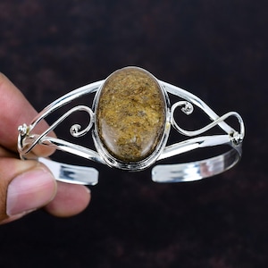 May include: A silver cuff bracelet featuring a large, oval-shaped, textured brown stone set in the center. The metal band is crafted with decorative swirling wire details on both sides of the central stone, creating an open, airy design.