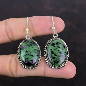 May include: A pair of silver earrings with green and black oval-shaped stones set in a silver frame. The stones have a mottled appearance.