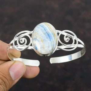 May include: A silver cuff bracelet featuring an oval-shaped gemstone with blue and white patterns. The bracelet has decorative swirls on each side of the gemstone.