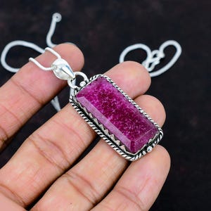 Op de afbeelding: Een rechthoekige hanger met een gefacetteerde, diep magentakleurige edelsteen in een zilverkleurig metalen frame met een gedraaide touwrand. De hanger hangt aan een dunne, zilverkleurige slangenketting met een gepolijst metalen oogje.