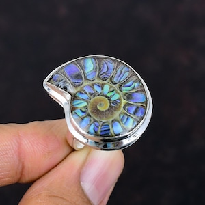Peut inclure: Bague en argent ornée d'un fossile d'ammonite poli. Le fossile présente un motif en spirale avec des reflets irisés bleus, verts et violets. La bague a un anneau simple en argent poli.