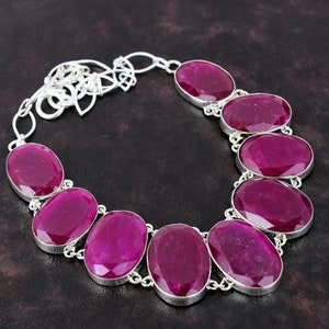 Può includere: Una collana d'argento con grandi pietre preziose ovali rosso scuro. Le pietre sono incastonate in castoni individuali d'argento e collegate da una delicata catena d'argento. La collana è mostrata su uno sfondo marrone scuro.