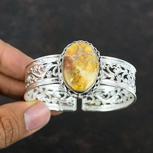 May include: A silver cuff bracelet with an oval-shaped yellow, brown, and white gemstone set in the center. The bracelet has a delicate, floral design.