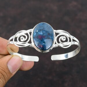 Könnte beinhalten: Ein silbernes Armreif-Armband mit einem ovalen blauen und rosafarbenen Edelstein, der in der Mitte eingesetzt ist. Das Armband hat ein zartes, filigranes Design mit geschwungenen Details.