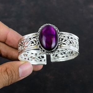 May include: A silver cuff bracelet featuring an oval-shaped, deep purple gemstone set in the center. The metal band displays an intricate, openwork filigree pattern of swirling vines and small flowers, with a twisted rope border around the stone.