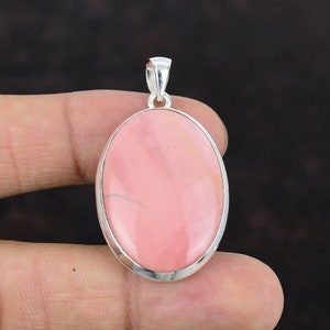 Pode incluir: Um pingente de prata com uma pedra oval rosa. O pingente é engastado em uma moldura de prata e tem um aro para prender uma corrente.