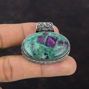 May include: Oval pendant featuring a green stone with vibrant magenta inclusions. The stone is set in a silver-toned metal with an ornate border and a decorative bail. The pendant is held by a hand against a dark background.