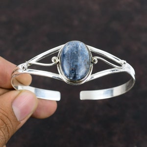 Könnte beinhalten: Ein silbernes Armreif-Armband mit einem blauen Edelstein, der in einer silbernen ovalen Fassung sitzt. Das Armband hat ein zartes, verziertes Design.