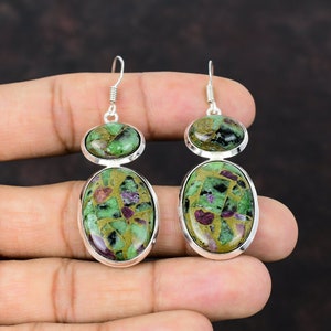 Op de afbeelding: Een paar zilveren oorbellen met groene en roze edelsteen druppels. De oorbellen hebben een dubbel-druppel ontwerp met een kleinere ronde steen aan de bovenkant en een grotere ovale steen aan de onderkant.