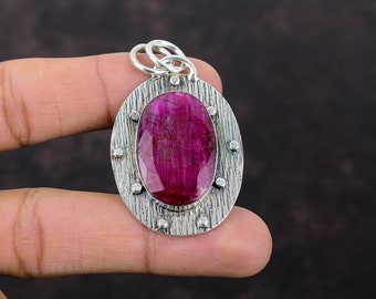 Può includere: Un ciondolo d'argento con una grande pietra di rubino ovale incastonata in una cornice d'argento testurizzata. Il ciondolo ha un'anellina per attaccare una catena.