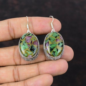 May include: Oval-shaped earrings with a silver-colored setting. The earrings feature a mosaic of green, purple, and gold-colored stones. The earrings are suspended from a simple hook.