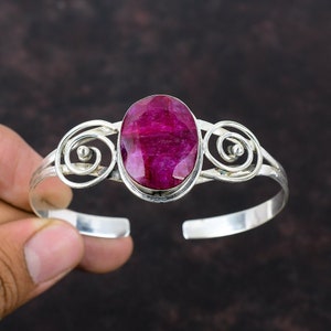 May include: A silver cuff bracelet featuring a large, oval-shaped, faceted deep pink ruby gemstone set in the center. The metal band is decorated with symmetrical silver wire scrollwork on both sides of the central stone setting.