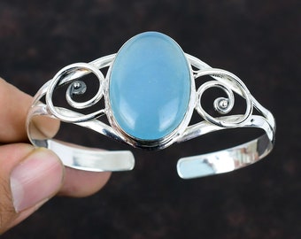 May include: A silver bangle bracelet with a light blue oval gemstone set in the center. The bracelet has a delicate, swirling design.