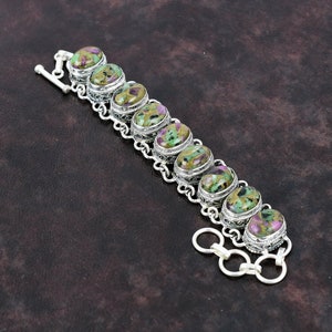 May include: A silver bracelet with a chain link design and oval-shaped green, brown, and pink gemstones. The bracelet has a toggle clasp.
