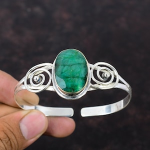 Puede incluir: Brazalete de plata con una gran esmeralda ovalada. La esmeralda está engastada en un bisel dorado y rodeada de espirales decorativas de plata. El brazalete tiene un diseño abierto.