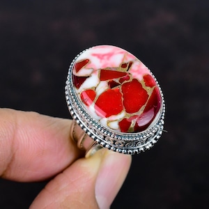 May include: Oval-shaped ring featuring a vibrant red, white, and gold gemstone. The gemstone is set in a silver band with intricate detailing around the edges. The ring's design showcases a unique blend of colors and textures, making it a striking piece of jewelry.