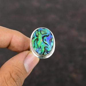 Pode incluir: Um anel de prata com uma incrustação oval de concha de abalone. A concha tem um padrão de redemoinho de verde, azul e roxo.