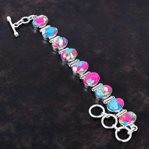 May include: A silver bracelet featuring oval-shaped stones in shades of pink, blue, and white. The bracelet has a toggle clasp closure and is displayed on a dark background. The stones are set in a silver metal setting.