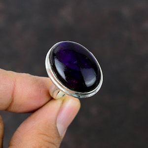 Puede incluir: Un anillo de plata con una gran piedra preciosa de amatista de forma ovalada. La piedra preciosa es de color morado oscuro y tiene un acabado liso y pulido.