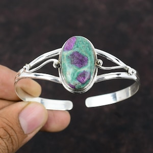May include: A silver bangle bracelet with a green and purple oval gemstone. The gemstone is set in a silver bezel and the bracelet has a delicate, curved design.