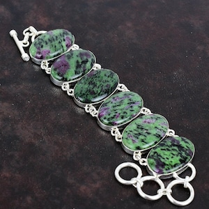 Könnte beinhalten: Ein silbernes Armband mit sieben ovalen, grün-violetten Edelsteinen. Das Armband hat einen Karabinerverschluss und ein Gliederketten-Design.