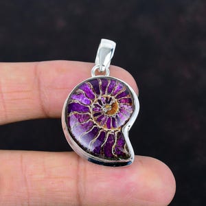 May include: A silver pendant featuring a polished, iridescent ammonite fossil. The fossil displays a spiral pattern with shades of purple, brown, and white. The pendant has a silver bail for attaching to a chain.