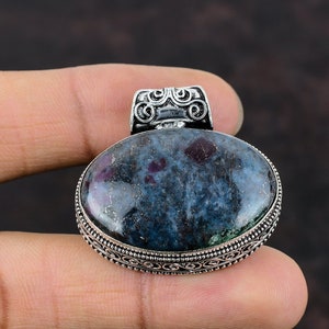 May include: Oval pendant featuring a blue-grey stone with red flecks, set in silver metal. The pendant has a decorative bail and a detailed silver border. The stone is approximately 3.8 cm long.