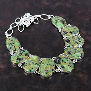 May include: A necklace featuring eight oval-shaped cabochons set in silver bezels. Each cabochon displays a mosaic pattern of green, black, and purple stone fragments held together by a gold-colored matrix. The stones are linked by a silver chain.
